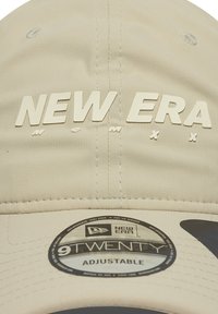 Beige New Era 9TWENTY állítható sapka kiemelkedő "NEW ERA" logóval és fekete swoosh-al a karimán.