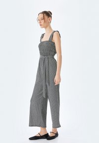 Svart och vitmönstrad jumpsuit med smockad byst, vida ben och knytbälte. Har volangaxlar och en avslappnad passform.