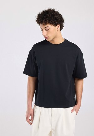 Junger Mann mit lockigem Haar, der ein locker sitzendes schwarzes T-Shirt mit kurzen Ärmeln und weiße Hose trägt, schaut nach unten, eine Hand in der Tasche.