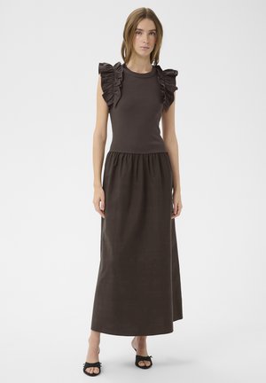 Femme portant une robe sans manches marron foncé avec des épaules à volants et une taille froncée, associée à des talons ouverts noirs, debout sur fond blanc.