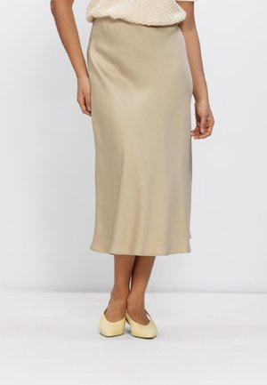 Woman wearing a beige midi-length satin skirt paired with cream-colored flat shoes, standing against a plain light background.