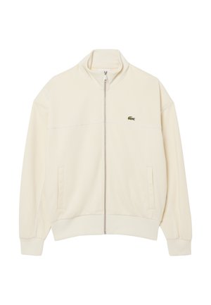 Chaqueta cremosa con cremallera, cuello alto, bolsillos laterales, puños y dobladillo acanalados, y pequeño logo de cocodrilo verde en el lado izquierdo del pecho.