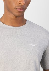 Tricou gri deschis, cu guler rotund, fabricat dintr-un amestec de bumbac, având un mic text alb cu logo-ul "Marc O'Polo DENIM" pe față.