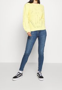 Pull en tricot jaune avec une coupe décontractée, présentant un motif texturé et un design à mailles ouvertes. Associé avec un jean slim bleu et des baskets noires.