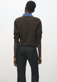 Persona con cabello corto, vistiendo un suéter marrón sobre una camisa azul con cuello y jeans oscuros, de pie contra una pared clara y lisa, de espaldas.