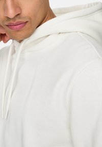 Haut à capuche blanc avec une texture douce, doté d'une capuche à cordons et de détails de couture visibles au niveau de l'encolure et des coutures d'épaule.