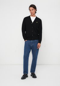 Black cardigan made of knitted fabric, featuring a V-neck, button closure, and a small yellow logo on the left chest. Paired with blue jeans.