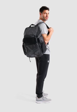 Zaino nero a scomparti multipli con superficie texturizzata, dettagli in rete e spallacci regolabili, indossato da una persona in abbigliamento sportivo grigio.