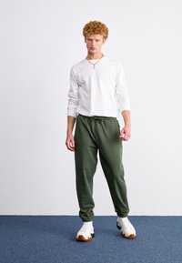 Camisa blanca de manga larga combinada con pantalones jogger verdes. Los joggers tienen una cinturilla elástica con cordón y tobillos ajustados. Zapatillas blancas con suela color beige.