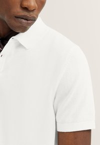 Weißes Poloshirt mit strukturiertem Strickmuster, kurzen Ärmeln und klassischem Kragen. Besitzt schwarze Knopfakzente an der Knopfleiste.