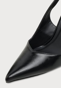 Gros plan d'une chaussure à talon haut noire brillante à bout pointu avec bride arrière sur fond uni.