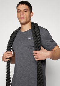T-shirt grigio a maniche corte con logo bianco, indossato da una persona che tiene delle robuste corde da allenamento nere, che mostra un design semplice e funzionale.