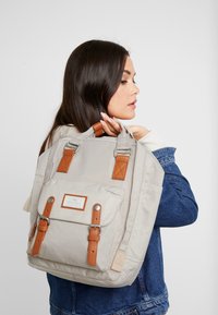 Sac à dos beige clair en tissu durable, avec des accents en cuir marron, une poche à rabat sur le devant et des bretelles réglables.