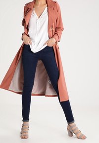 Kvinna klädd i en rostfärgad trenchcoat, vit blus, mörka skinny jeans och grå remsandaler med klack, står med händerna i fickorna.