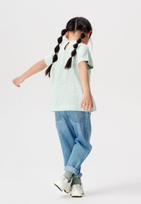 Camicia verde chiaro a maniche corte con dettaglio a taglio sul retro, abbinata a jeans in denim blu larghi e sneakers bianche con dettagli verdi.