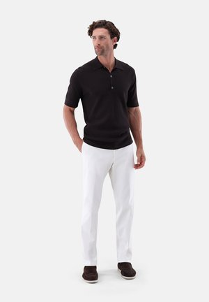 Mann steht mit Händen in den Taschen, trägt ein schwarzes kurzärmeliges Polo, weiße Hose und dunkelbraune Schuhe vor einfarbigem Hintergrund.