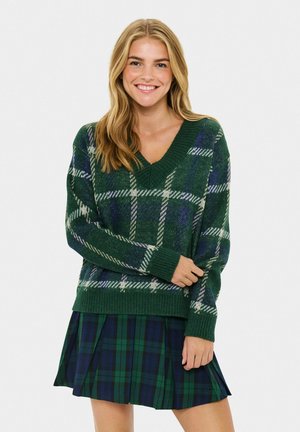 Smiling woman with long blonde hair wearing a green and blue plaid V-neck sweater and a matching pleated skirt.