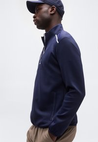 Άνδρας που φοράει μπλε-σκούρο ζακέτα με φερμουάρ και καπέλο που ταιριάζει, με ανοιχτόχρωμο παντελόνι, στέκεται σε προφίλ μπροστά σε απλό λευκό φόντο.