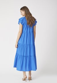 Rochie maxi albastră cu mâneci scurte volan, design în straturi, talie elastică și fustă fluidă; purtată cu sandale cu toc transparent.