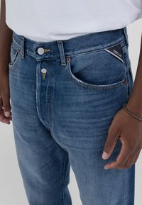 Jean en denim dans un bleu moyen avec un léger effet usé. Doté d'un design classique �à cinq poches, d'un bouton en métal et d'une étiquette avec logo sur la ceinture.