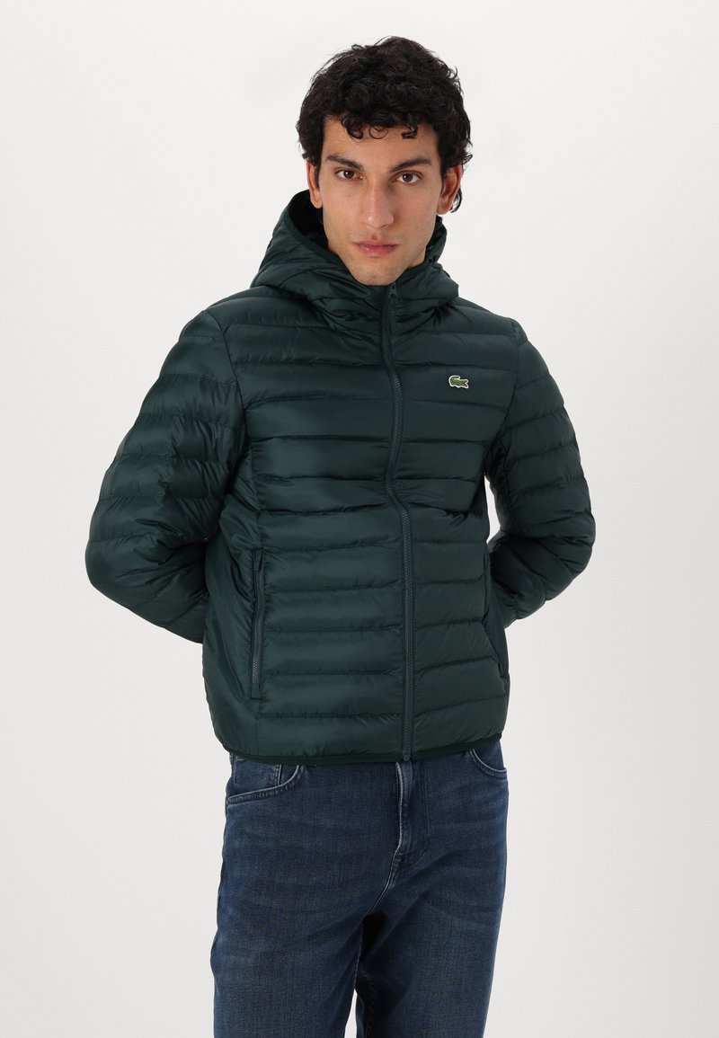 Chaqueta verde acolchada con capucha, con costuras horizontales, bolsillos frontales y un logo en el pecho. Lleva puesta sobre unos jeans azules.