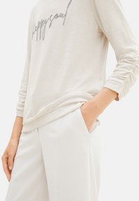 Helles beige langärmeliges Oberteil mit "happysoul" in silberner Stickerei. Weicher Stoff mit relaxter Passform, kombiniert mit weißen Slim-Fit-Hosen.