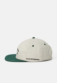 Casquette avec un corps beige et un visière verte. Présente un texte brodé sur le côté et un bouton vert sur le dessus. Fabriquée en tissu.