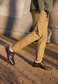 Chaussures habillées noires en cuir avec une finition lisse, pantalon beige sur mesure, marchant sur une surface en pavés texturée, ombre projetée par la lumière au-dessus.