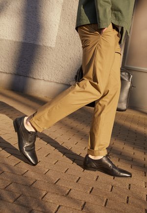 Chaussures habillées noires en cuir avec une finition lisse, pantalon beige sur mesure, marchant sur une surface en pavés texturée, ombre projetée par la lumière au-dessus.