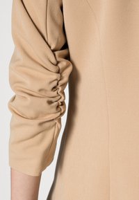 Gros plan d'une manche beige avec des détails de tissu froncé le long du bras, montrant la texture et les coutures sur un matériau tissé lisse.