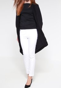 Svart öppenframs cardigan, lätt material, långa ärmar, kombinerad med en figurnära svart topp och vita smala byxor, avslutad med svarta klackar.