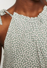 Haut à motif floral avec un fond vert clair, encolure à volants et bretelles réglables à nouer. Le tissu semble doux et léger.