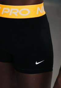 Μαύρα αθλητικά σορτς με κίτρινη μέση που γράφει "NIKE PRO" και λευκό σήμα Nike στο κάτω δεξί πόδι, φορεμένα από ένα άτομο.