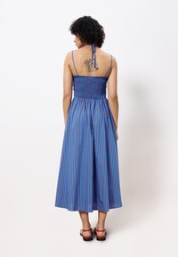 Blau-rot gestreiftes Midikleid mit gerafftem Oberteil, Neckholder und fließendem Rock. Das Modell trägt rote Sandalen und zeigt ein Tattoo auf ihrem Rücken.