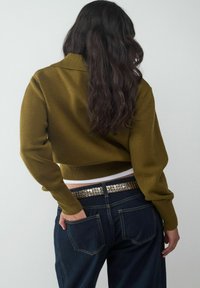 Olivgrüner, kurzer Strickpullover mit Rippenkragen, kombiniert mit dunkelblauen Jeans, die einen metallisch genagelten Gürtel und Gesäßtaschen haben.
