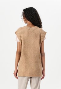 Tan strikket vest med vertikale ribber, ermeløs design og rund utringning, brukt over en lys rosa skjorte.