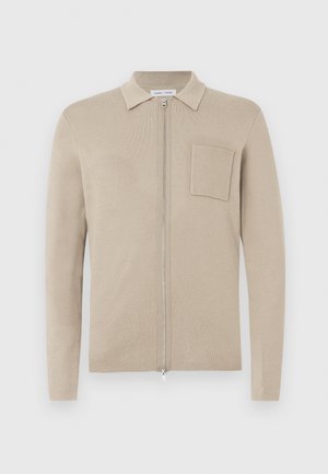 Cardigan beige con zip, maniche lunghe, colletto e tasca frontale. Realizzato in morbido materiale a maglia, presenta una texture liscia e un design minimalista.