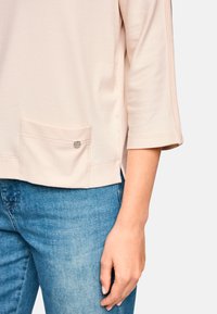 Pale beige top with three-quarter sleeves, featuring a front pocket and metal accent. Worn with light blue denim jeans. Soft texture.