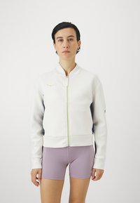 Hvit zip-up jakke med kontrasterende mørkeblå sider, grønn glidelåsdetalj, ribbestrikkede mansjetter og en liten gull-logo på brystet. Brukt med lilla shorts.