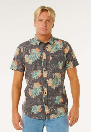 Jeune homme debout, portant une chemise grise à manches courtes avec de grands motifs floraux orange et bleu sarcelle, ainsi qu'un jean bleu clair.