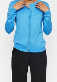 Ljusblå cardigan i mjukt tyg, med knäppning framtill, ribbade ärmslut och en figurnära design, kombinerad med svarta byxor.