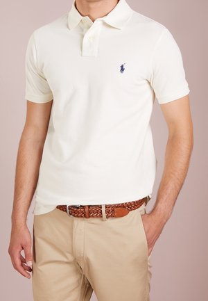 Mężczyzna w białej koszulce polo z małym granatowym logo, w beżowych spodniach i brązowym plecionym skórzanym pasku, stoi z jedną ręką w kieszeni.