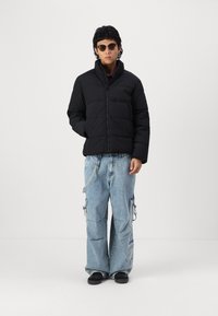 Black padded jacket with high collar and zipper, paired with light blue wide-leg cargo jeans featuring pockets and loops. Black shoes and sunglasses.