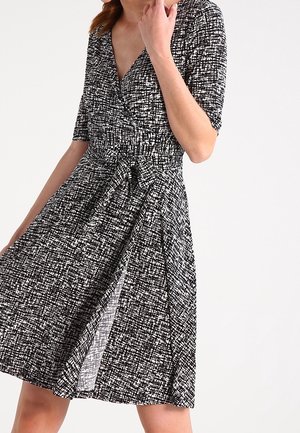 Femme portant une robe portefeuille à motif noir et blanc avec des manches trois-quarts, nouée à la taille, posant devant un fond uni.