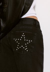 Čierne denimové džínsy so vzorom hviezd vytvoreným zo strieborných cvokov na zadnej vrecku a koženou nášivkou značky na páse.