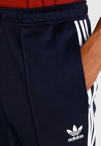 Námořnické modré sportovní kalhoty vyrobené z hladkého materiálu, s bílými bočními pruhy, logem Adidas a elastickým pasem se šňůrkou.
