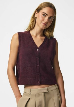 Femme aux cheveux châtain clair porte un gilet sans manches violet foncé boutonné et un pantalon beige plissé taille haute sur un fond blanc uni.