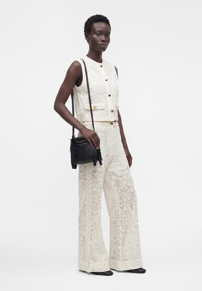 Femme portant un gilet blanc brodé sans manches et un pantalon large assorti, tenant un sac bandoulière noir à franges, debout sur fond blanc.