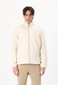 Cream fleece jacket with a high collar, front zipper, and side pockets. Features subtle branding on the chest and a relaxed fit.