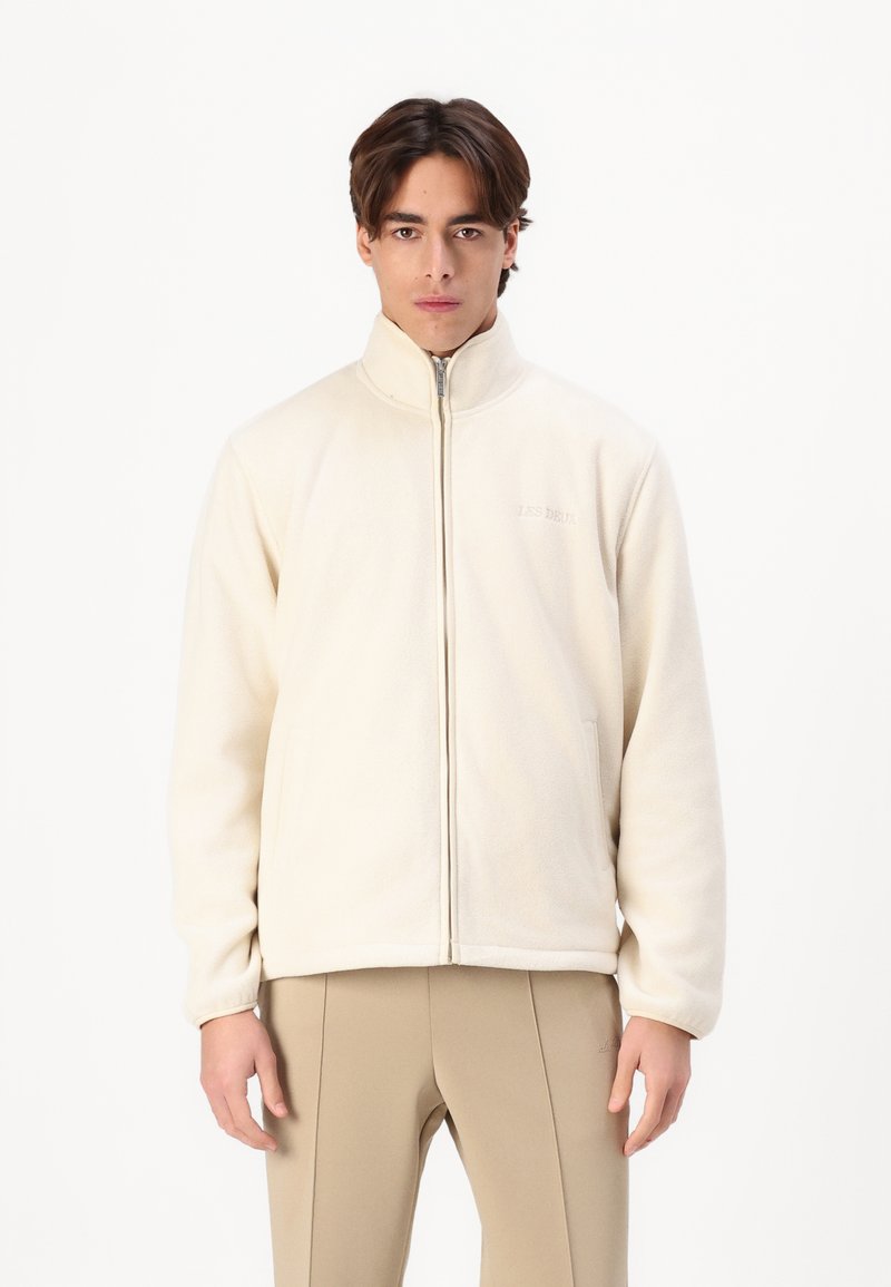 Cream fleece jacket with a high collar, front zipper, and side pockets. Features subtle branding on the chest and a relaxed fit.